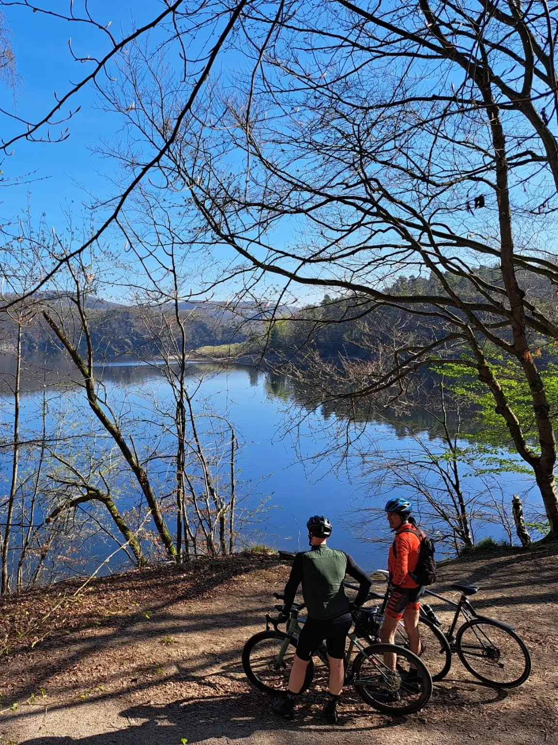 Twee fietsers kijken tussen kale bomen uit over een stil stuwmeer